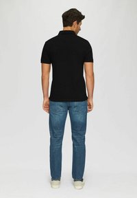 Schwarzes Poloshirt mit kurzen Ärmeln und einem Standardkragen, kombiniert mit blauen Jeans und hellen Turnschuhen. Von hinten gesehen.