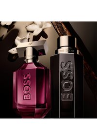 Twee BOSS The Scent parfumflessen, één dieproze met een zilveren dop, één zwart met een zilveren dop, omringd door witte bloemen en een vanilestokje.