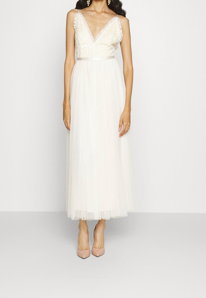 Femme portant une robe midi blanche sans manches avec un corsage en dentelle à col en V et une jupe en tulle, associée à des escarpins pointus couleur nude.