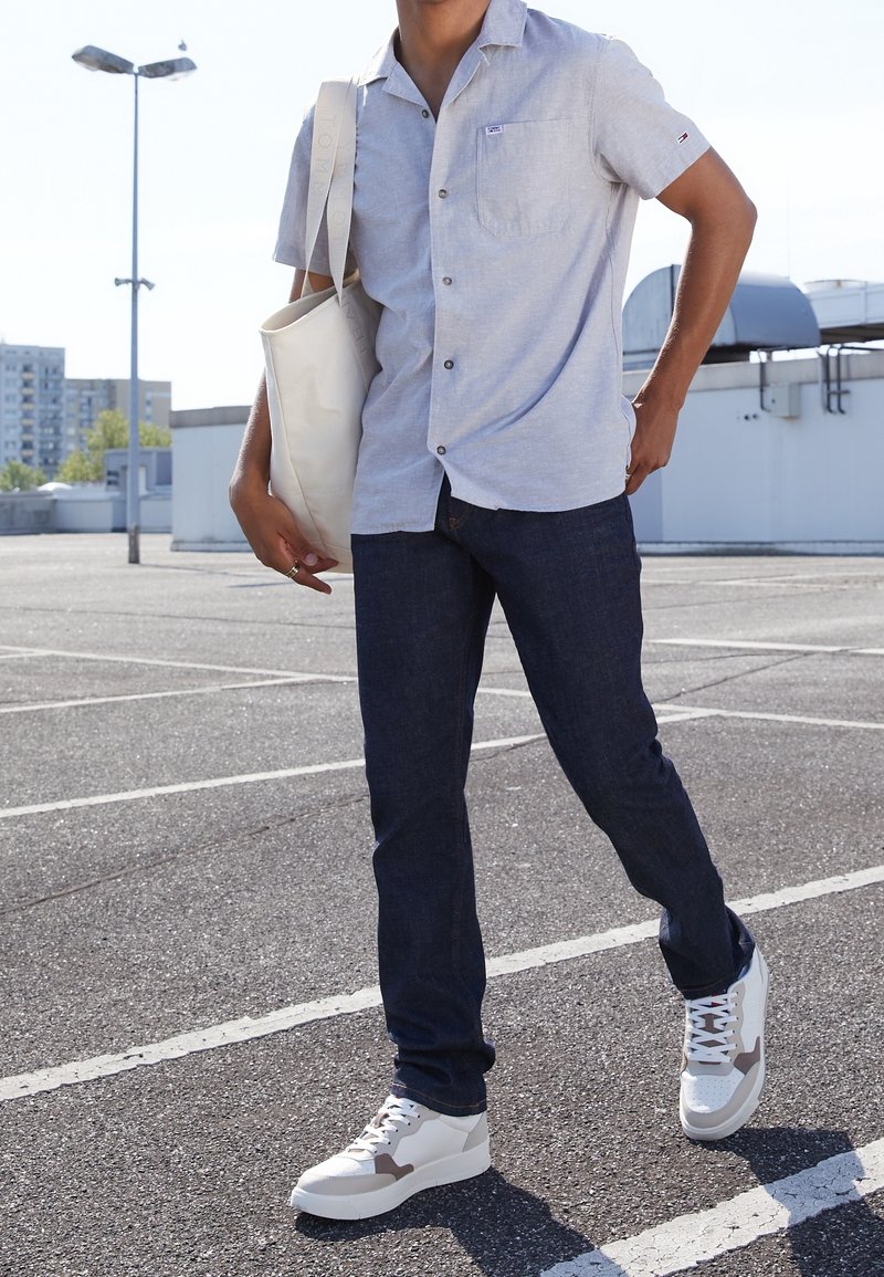 Homme portant une chemise à manches courtes bleu clair, un jean foncé, des baskets blanches et portant un sac fourre-tout crème dans un parking extérieur.