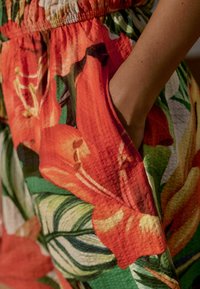Bunt bedruckter Stoff mit großen orangefarbenen und roten Blumen, grünen Blättern und einer strukturierten Oberfläche; umfasst einen elastischen Bund mit Taschen.