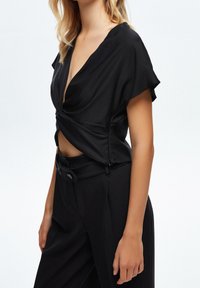 Černá blůza s V-výstřihem a krátkými rukávy, s předním designem ve tvaru twist. Nosená s černými kalhotami s vysokým pasem. Hladká textilie.