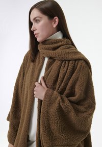 Abrigo de fleece marrón y texturizado que se envuelve alrededor de los hombros, con un alto cuello y un material grueso y suave, además de un corte holgado.