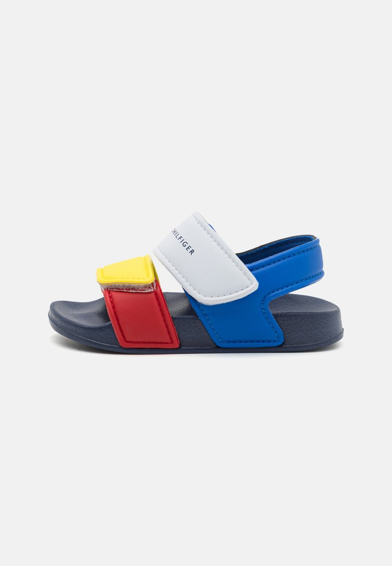 Children's sandals with a blue, red, and white design. Features adjustable Velcro straps and a textured rubber sole. Yellow accent square.