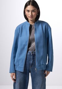 Ljusblå zip-up jacka med hög krage, tillverkad av mjukt tyg. Har sidofickor och en avslappnad passform, i kombination med en mörk texturerad topp.