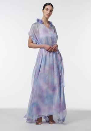 Mujer de pie con un vestido largo y fluido de acuarela en tonos pastel morado y azul, con tela translúcida, mangas con volantes y detalles florales en el cuello.