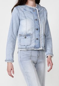 Light blue denim jacket with braided trim, frayed edges, five buttons, two front pockets, and a round collar. Smooth texture throughout.