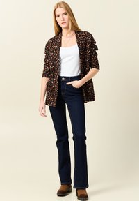 Zwarte cardigan met bloemenpatroon over een witte tanktop, gecombineerd met donkere flared jeans en bruine hoge laarzen, tegen een neutrale achtergrond.