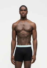 Αθλητικός άνδρας με σκούρο δέρμα, όρθιος και στραμμένος προς τα εμπρός, φοράει μαύρα μπόξερ Björn Borg με ανοιχτό μπλε ζωνάκι στη μέση.