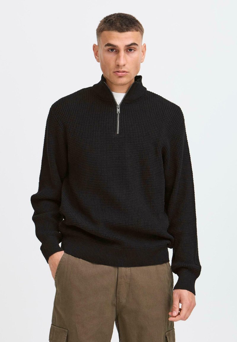 Schwarzer Strickzippullover mit Rippbündchen und -saum, der ein strukturiertes Muster und einen kurzen Reißverschluss am Kragen aufweist. Getragen mit Cargo-Hosen.