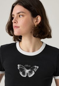 Μαύρο μπλουζάκι με λευκή ρίγα στο γιακά. Διαθέτει γκρι εκτύπωση με πεταλούδα στο κέντρο και το κείμενο "Levi's" από πάνω. Λείο ύφασμα, εφαρμοστό σχέδιο.