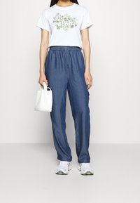 Mujer vestida con una camiseta blanca de logo floral, pantalones azules con cordón, zapatillas blancas, sosteniendo un pequeño bolso blanco, fondo claro y liso.