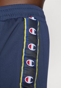 Kék sportnadrág elasztikus derékrésszel, az oldalsó panelen multicolór Champion logókkal piros, kék és sárga színekben.