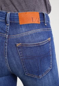 Jeans i denim i djupblått med en orange solbrun läderlap med texten "T/J." Har standard bakfickor med kontrasterande sömnadsdetaljer.