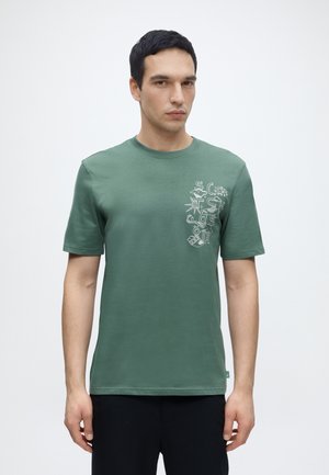 Homme portant un t-shirt vert à manches courtes avec des lettres graphiques abstraites blanches sur la poitrine gauche, debout devant un fond blanc uni.