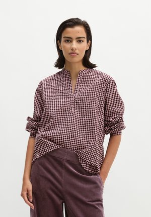 Femme aux cheveux courts et foncés portant un chemisier rose à pois noirs à manches longues et un pantalon en velours côtelé violet, une main dans la poche.
