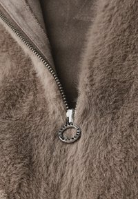 Brown faux fur jacket with a silver zipper. The material has a soft, textured appearance and a smooth interior lining. Zipper pull features a circular design.