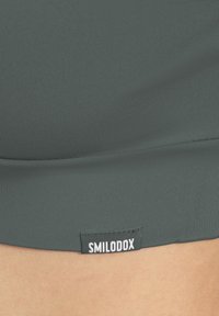Dunkelgrünes, tailliertes Oberteil aus strukturiertem Stoff, mit einem kleinen weißen "SMILODOX"-Etikett nahe dem Saum. Das Design ist nahtlos und schlank.