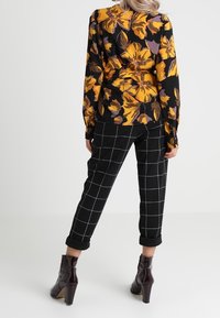 Svart blommig blus med gula och lila blommor, parat med svarta rutiga byxor och glänsande ankelskor.