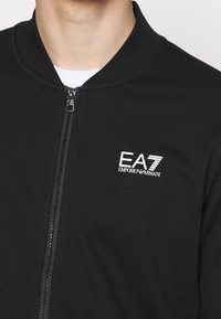 Veste noire zippée en tissu doux, avec col montant, fermeture éclair latérale et logo blanc EA7 Emporio Armani. Design minimaliste sans motifs.
