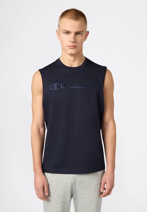 Jeune homme aux cheveux blonds courts portant un débardeur bleu marine Champion et un pantalon de survêtement gris clair, debout devant un fond blanc.