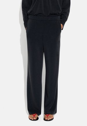 Osoba ubrana w luźne czarne spodnie z elastycznym pasem i czerwone sandały na palce, ręce w kieszeniach, stojąca na białym tle.