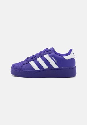 Sneaker Adidas Superstar viola con strisce bianche, linguetta sul tallone bianca e punta a conchiglia testurizzata su uno sfondo chiaro uniforme.