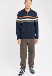 Polo à manches longues bleu marine avec des rayures orange, blanches et olives sur la poitrine, associé à un pantalon cargo gris et des baskets bleu marine.