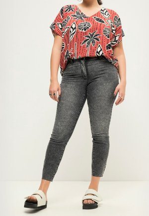 Femme portant un haut corail à manches courtes avec un imprimé floral noir et blanc, un jean skinny taille haute délavé acide, et des sandales blanches à enfiler.