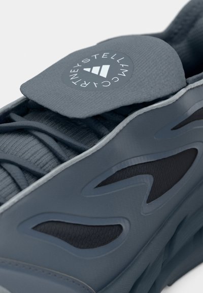 Γκρι αθλητικό παπούτσι με πολυεπίπεδο σχέδιο, διαθέτει υφασμάτινο επάνω μέρος με υφή, λεία ελαστική σόλα και λογότυπο στη γλώσσα. Διακρίνονται χαρακτηριστικές οπές αερισμού.