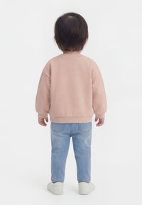 Tout-petit debout de dos, portant un sweat-shirt rose clair, un jean bleu et des baskets blanches sur un fond blanc.