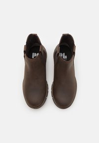 Des bottines en cuir marron avec des panneaux élastiques sur les côtés, des bouts ronds et des semelles en caoutchouc texturé. L'intérieur présente une semelle noire avec logo.