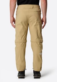 Beige ulkopantit, joissa on vetoketjullinen alaosa, kevyt kangas ja joustava vyötärö. Oikeassa lahkeessa on logo.