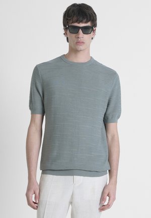Maglione a maniche corte in maglia testurizzata di teal tenue con righe orizzontali; colletto rotondo e orlo a costine; indossato con pantaloni chiari.