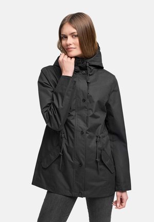 Žena oblečená v čiernej kapucňovej bunde so zapínaním na gombíky a sťahovacou šnúrkou, pózujúca s rukou pri brade na jednofarebnom pozadí.