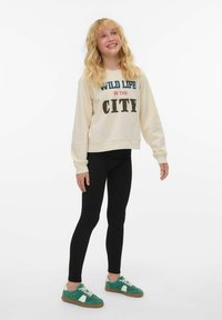 Sweatshirt crème avec le texte "VIE SAUVAGE EN VILLE", leggings noirs et baskets vertes avec des accents blancs. Style décontracté, coupe relax.