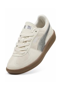 Zapatilla blanca con materiales de ante y sintéticos, logo PUMA plateado, detalle gris texturizado y suela de goma marrón. Cuenta con cordones planos.