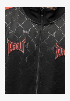 Melns zipa tīkla jaka ar dimanta rakstu, kurai krūtīs un plecos ir sarkanie "TAPOUT" logotipi. Izgatavota no gludas, vieglas auduma.