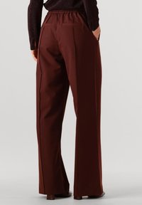 Pantalon large marron avec taille élastique et deux poches arrière, présentant un passepoil noir le long des coutures latérales. Texture de tissu lisse.