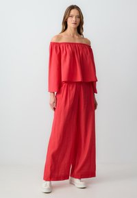 Rotes schulterfreies Oberteil mit rüschengesäumtem Saum, kombiniert mit weit geschnittenen Hosen. Strukturiertem Stoff, lässiger Schnitt und weißen Sneakers.