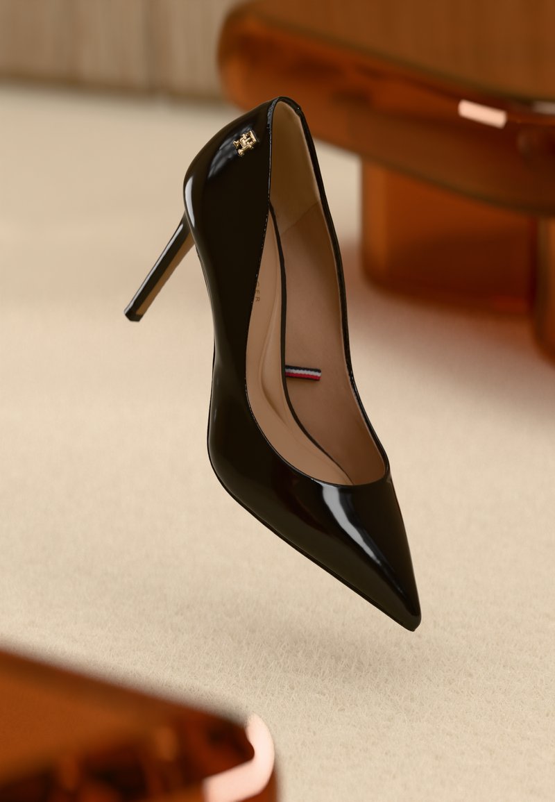 Chaussure à talon aigu en cuir verni noir avec un nez pointu et un emblème doré près de l'ouverture, sur un fond texturé beige.