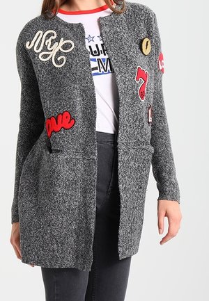 Cardigan gris à ouverture devant avec des patchs rouges, blancs et jaunes comprenant "NYC", "love", "7" et un éclair, porté sur un t-shirt graphique blanc.