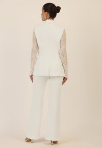 White tailored suit with a solid silhouette and sheer lace long sleeves. Flared pants complete the ensemble, featuring intricate lace detailing.