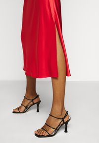 Robe midi en satin rouge avec une fente sur le côté, accompagnée de sandales à talons noires à lanières. Texture lisse, design ajusté.
