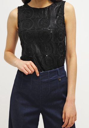 Femme portant un haut sans manches en dentelle noire avec des motifs circulaires et un pantalon large taille haute bleu foncé, sur un fond uni.