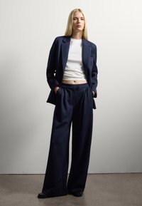 Completo elegante navy con giacca struturata e pantaloni a gamba larga, abbinato a un top bianco aderente, su uno sfondo testurizzato.