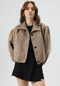 Beige suede jacket with a collar, front buttons, and voluminous sleeves. Worn over a black top and skirt. Simple, functional design.