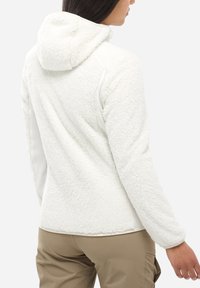 Kobieta ubrana w białą puszystą kurtkę z kapturem i beżowe spodnie, pokazana od tyłu i lekko obrócona na bok.