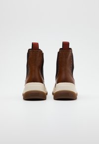 Botas de piel marrón hasta el tobillo con paneles elásticos laterales, entresuela blanca, suela de goma y tiradores rojos en el talón. Acentos texturizados visibles.