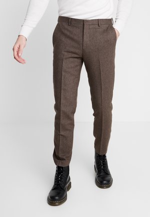 Man in een bruine op maat gemaakte broek en zwarte veterschoenen, staand met één hand in zijn zak tegen een witte achtergrond.